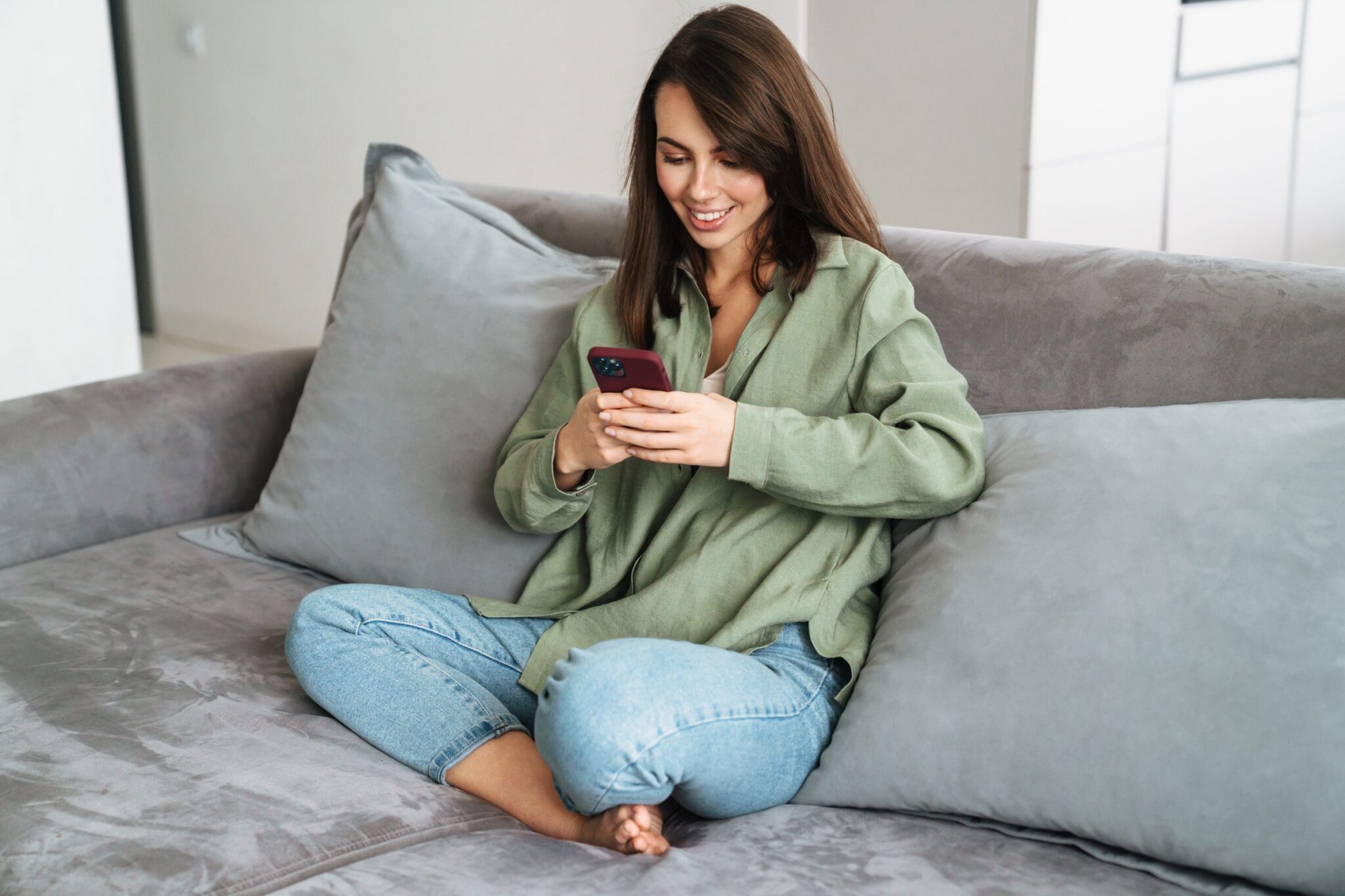Junge Frau sitzt entspannt auf dem Sofa und schaut lächelnd auf ihr Handy